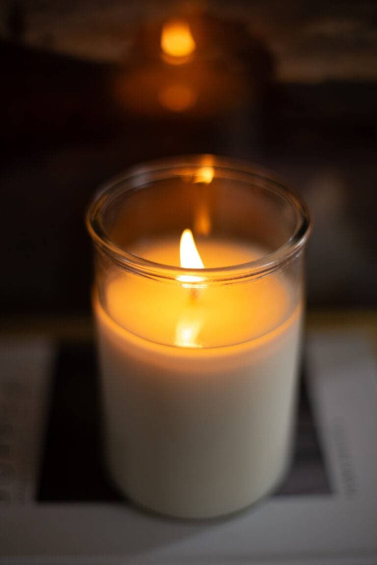 A lit white candle in a glass jar, emitting a warm glow and creating a calm, cozy atmosphere.